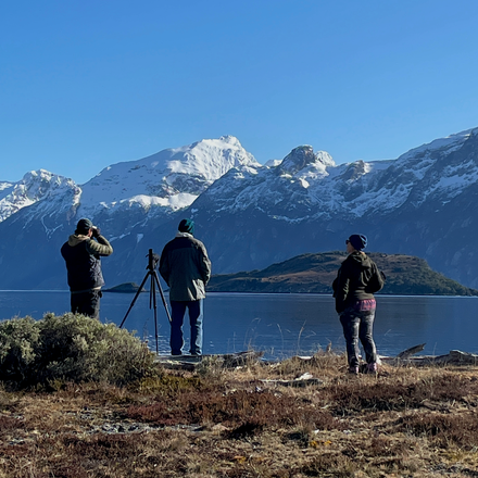 Three birdwatchers beside a lake with snow-covered mountains beyond