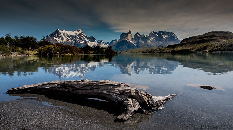 The mountains of Torres del Paine reflected in a lake, with driftwood on the shore